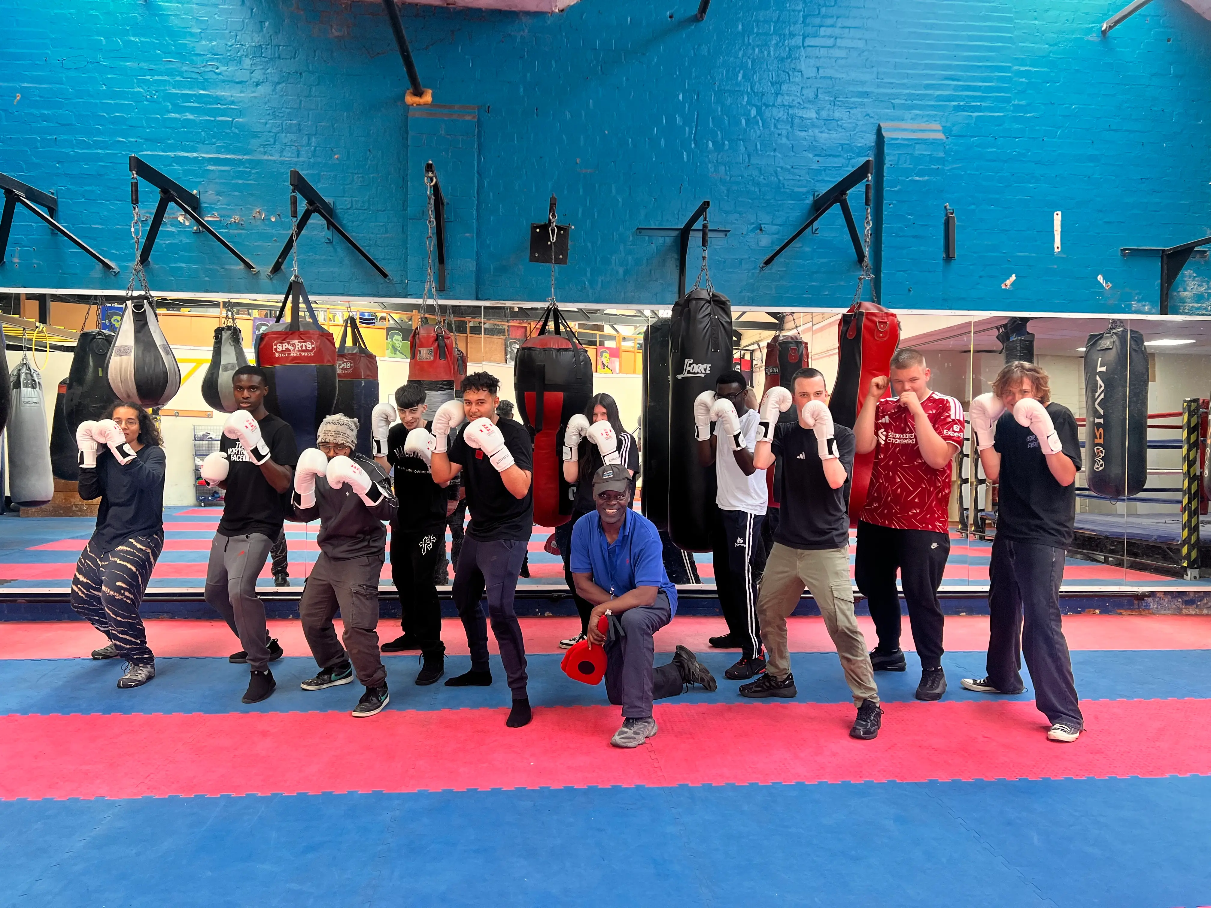 Members of a recreational boxing session standing in stance with gloves raised, a coach kneeling among them, in the Waterfront gym